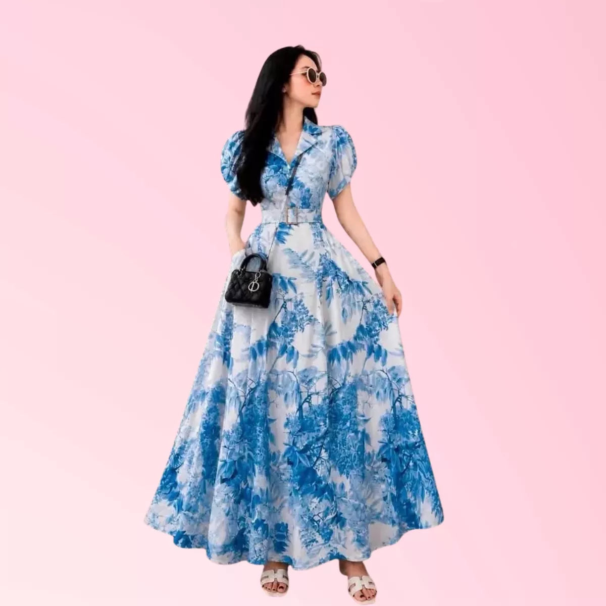 A woman modeling a blue and white floral print maxi dress with puff sleeves, a collared neckline, and a matching belt, styled with a small black handbag and sunglasses against a solid pink background.