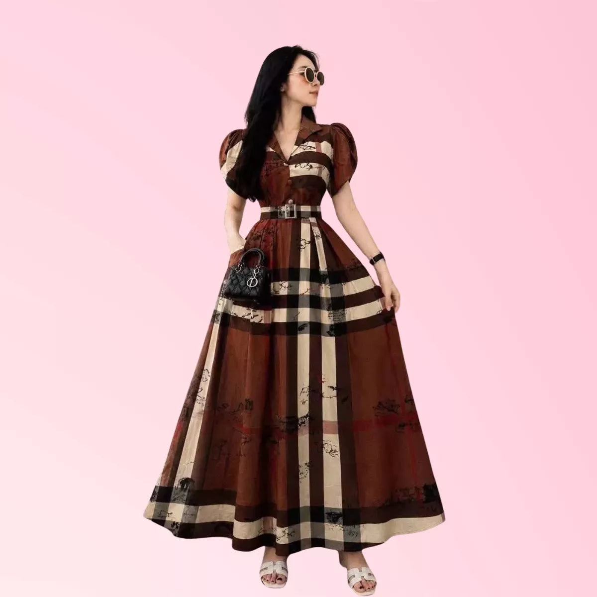 A woman modeling a brown and beige plaid maxi dress with puff sleeves, a belted waist, and a full skirt, set against a solid pink background.