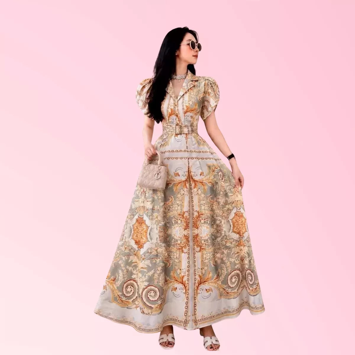 A woman modeling a long, puff-sleeve maxi dress with an ornate baroque-style print in beige, gold, and brown tones, set against a solid light pink background.